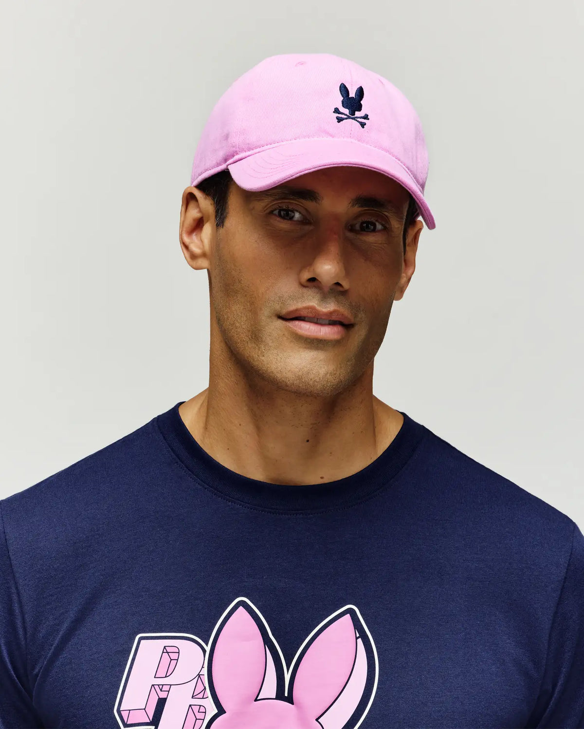 Jimmy Baseball Cap in pink with a navy embroidered Psycho Bunny skull logo, paired with a navy jersey t-shirt featuring a large, central pink 3D Psycho Bunny graphic print.