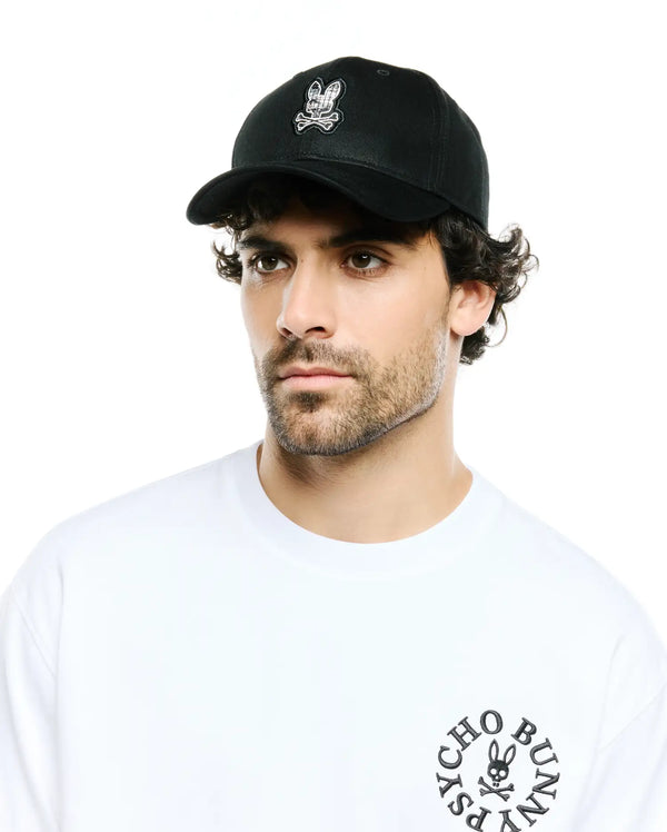 Men's Alfie Baseball Cap in black with a front embroidered white and black plaid bunny skull logo, paired with a white jersey t-shirt featuring a black circular Psycho Bunny text and skull logo on the left chest.