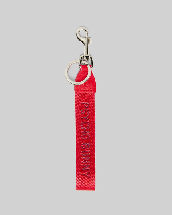 The PSYCHO BUNNY WOVEN TAPE KEYCHAIN - MO1700001 by Psycho Bunny features "PSYCHO BUNNY" in purple on red fabric, with a logo-etched keyring and lobster clasp, shown against a light gray background.