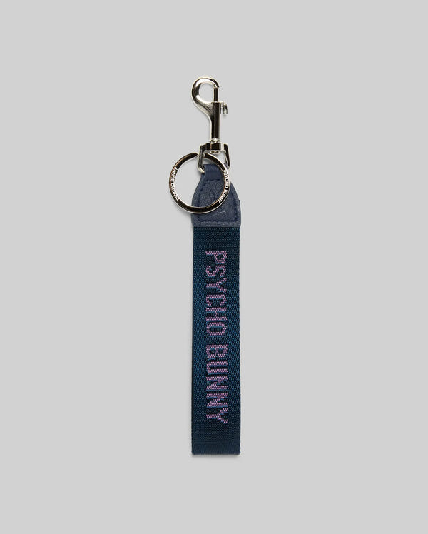 The PSYCHO BUNNY WOVEN TAPE KEYCHAIN - MO1700001 by Psycho Bunny features sleek blue fabric, a silver metal ring and clasp, and "PSYCHO BUNNY" embroidered in light purple on the strap, shown against a gray background for a stylish look.