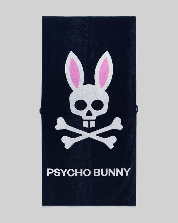 The Psycho Bunny MENS CLASSIC BUNNY TOWEL - MO1700004 is a navy blue towel with a white skull and crossbones, pink bunny ears, and "PSYCHO BUNNY" in bold white letters on a gray background.