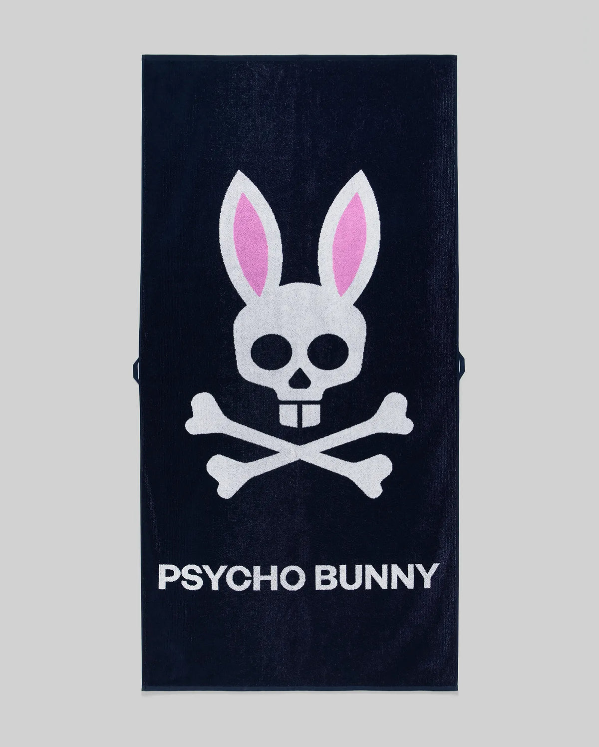 The Psycho Bunny MENS CLASSIC BUNNY TOWEL - MO1700004 is a navy blue towel with a white skull and crossbones, pink bunny ears, and "PSYCHO BUNNY" in bold white letters on a gray background.