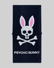 The Psycho Bunny MENS CLASSIC BUNNY TOWEL - MO1700004 is a navy blue towel with a white skull and crossbones, pink bunny ears, and "PSYCHO BUNNY" in bold white letters on a gray background.