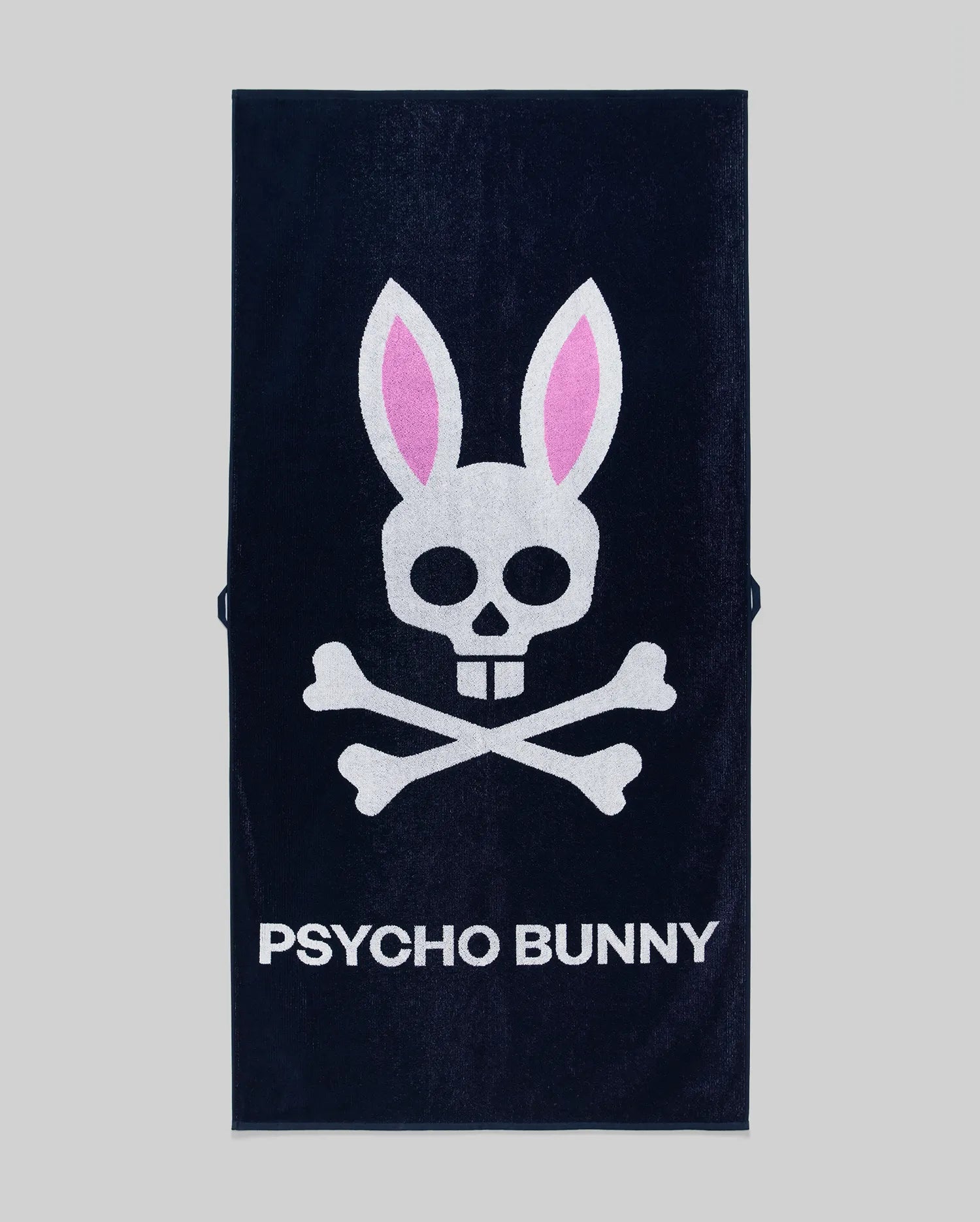 The Psycho Bunny MENS CLASSIC BUNNY TOWEL - MO1700004 is a navy blue towel with a white skull and crossbones, pink bunny ears, and "PSYCHO BUNNY" in bold white letters on a gray background.