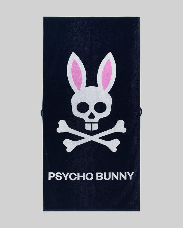 The Psycho Bunny MENS CLASSIC BUNNY TOWEL - MO1700004 is a navy blue towel with a white skull and crossbones, pink bunny ears, and "PSYCHO BUNNY" in bold white letters on a gray background.