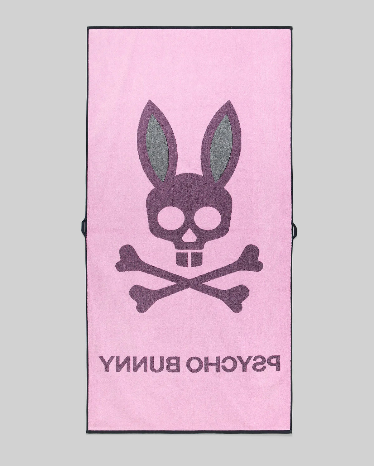The MENS CLASSIC BUNNY TOWEL - MO1700004 by Psycho Bunny is a pink rectangular towel featuring a bold skull and crossbones with bunny ears, plus "PSYCHO BUNNY" in large text beneath the design.