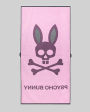 The MENS CLASSIC BUNNY TOWEL - MO1700004 by Psycho Bunny is a pink rectangular towel featuring a bold skull and crossbones with bunny ears, plus "PSYCHO BUNNY" in large text beneath the design.