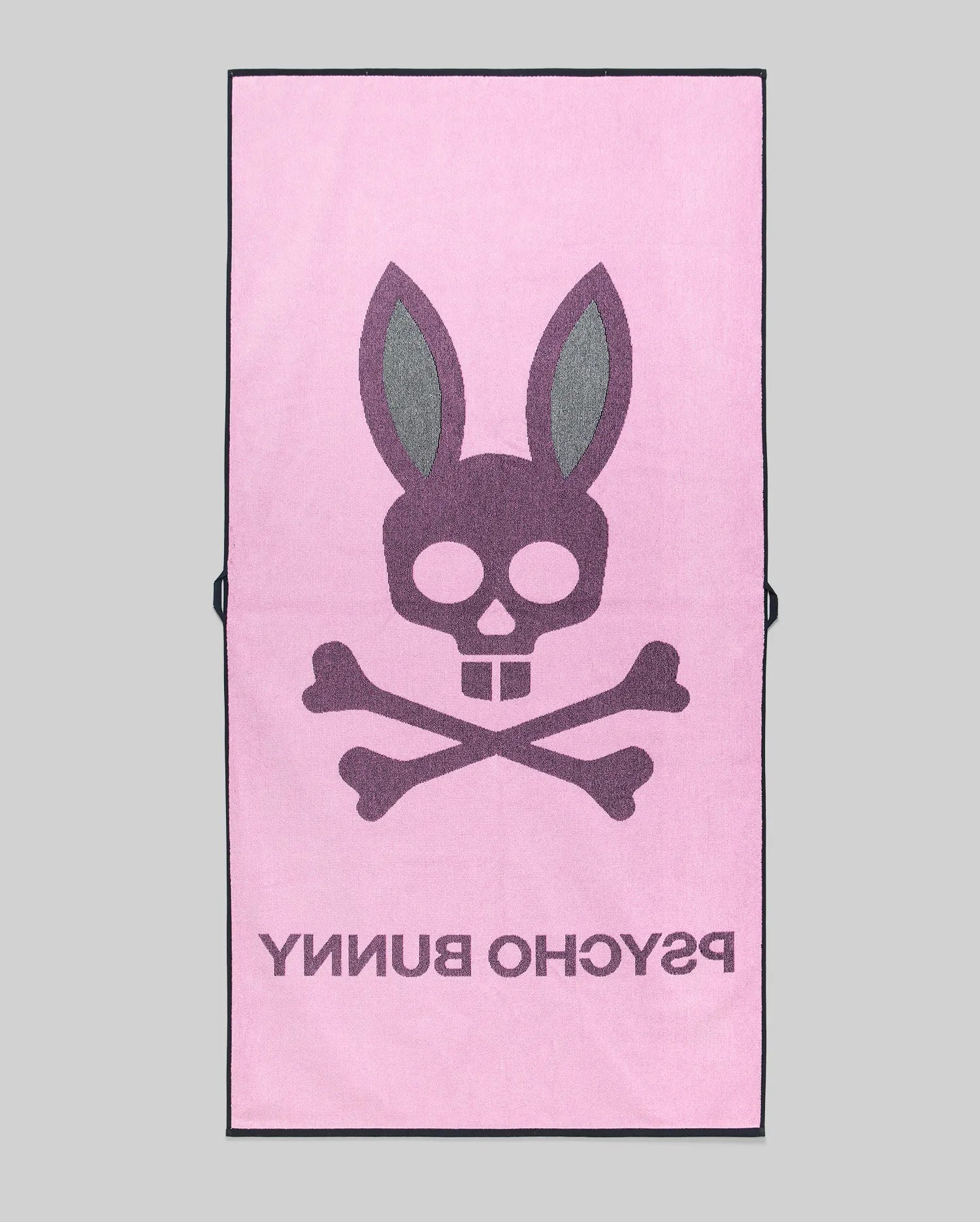 The MENS CLASSIC BUNNY TOWEL - MO1700004 by Psycho Bunny is a pink rectangular towel featuring a bold skull and crossbones with bunny ears, plus "PSYCHO BUNNY" in large text beneath the design.