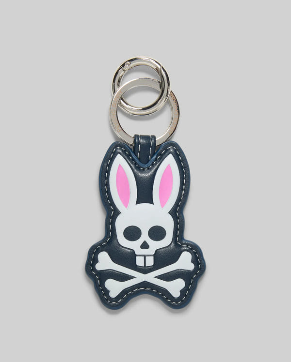 The Psycho Bunny MENS PUFFER BUNNY KEYCHAIN (MO1700008) features a cartoon bunny with pink ears and a white skull-and-crossbones face on a dark background, plus two silver key rings—an eye-catching addition to any men’s keychain collection.