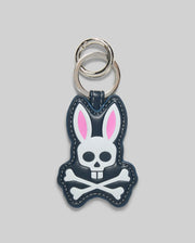 The Psycho Bunny MENS PUFFER BUNNY KEYCHAIN (MO1700008) features a cartoon bunny with pink ears and a white skull-and-crossbones face on a dark background, plus two silver key rings—an eye-catching addition to any men’s keychain collection.