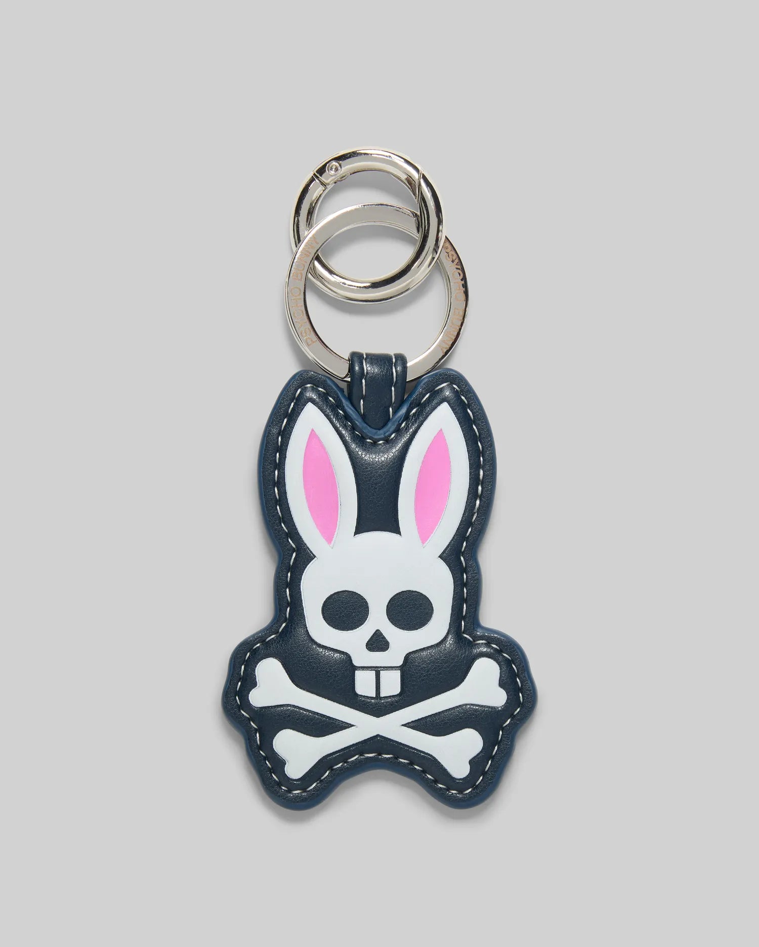 The Psycho Bunny MENS PUFFER BUNNY KEYCHAIN (MO1700008) features a cartoon bunny with pink ears and a white skull-and-crossbones face on a dark background, plus two silver key rings—an eye-catching addition to any men’s keychain collection.