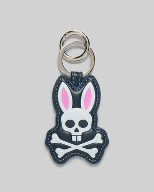 The Psycho Bunny MENS PUFFER BUNNY KEYCHAIN (MO1700008) features a cartoon bunny with pink ears and a white skull-and-crossbones face on a dark background, plus two silver key rings—an eye-catching addition to any men’s keychain collection.