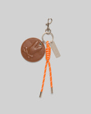 The Psycho Bunny DISC KEYCHAIN - MO1700013 features a round faux-leather tag with an embossed bird, orange knotted cord with metal tips, silver tag, and lanyard-clasp, displayed on a light gray background.