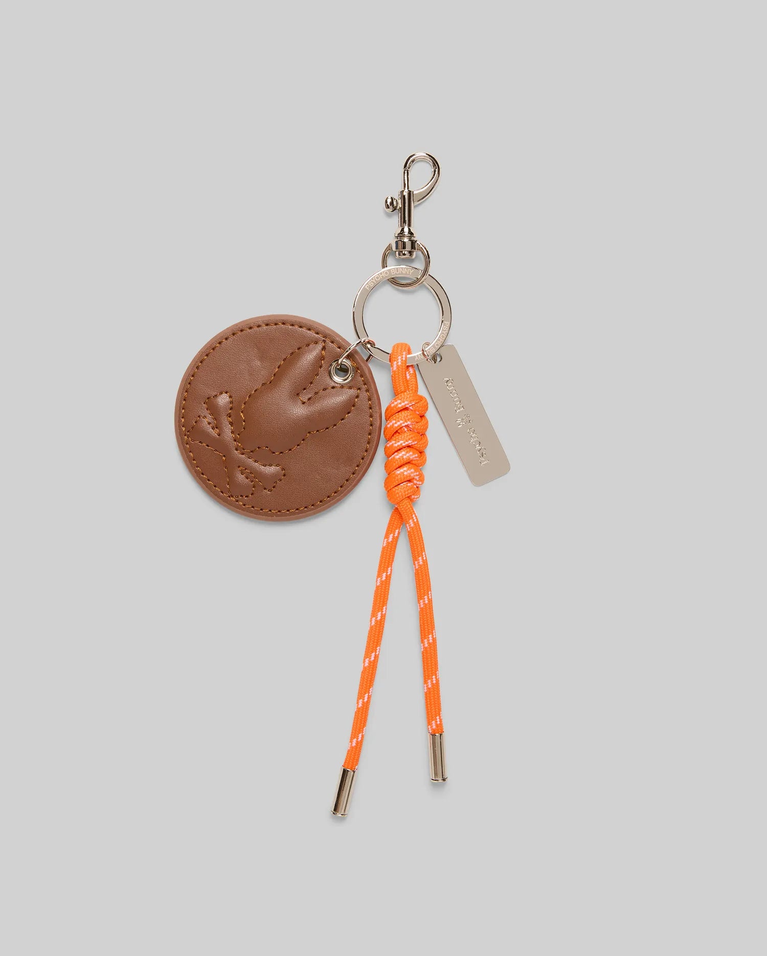 The Psycho Bunny DISC KEYCHAIN - MO1700013 features a round faux-leather tag with an embossed bird, orange knotted cord with metal tips, silver tag, and lanyard-clasp, displayed on a light gray background.