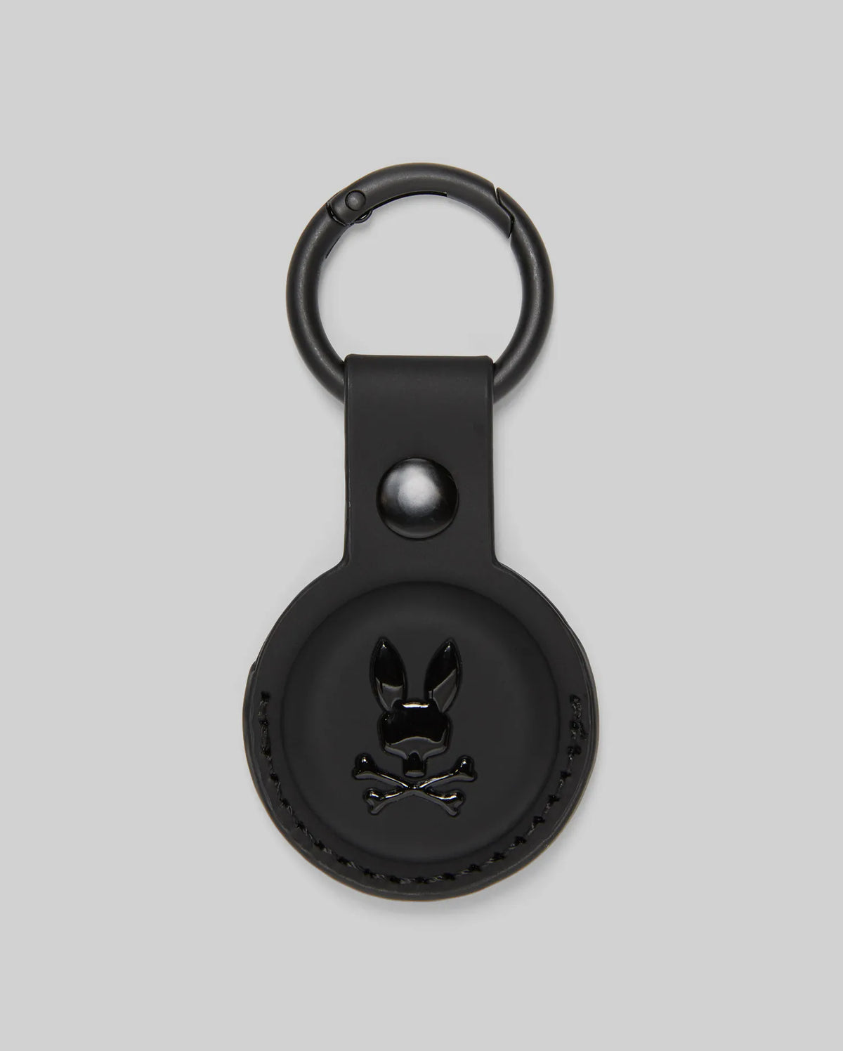 The Psycho Bunny MENS AIR TAG HOLDER - MO1700017 is a round, black leather holder with an embossed bunny logo, snap button, and metal keyring—ideal as a stylish luggage tag.