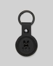 The Psycho Bunny MENS AIR TAG HOLDER - MO1700017 is a round, black leather holder with an embossed bunny logo, snap button, and metal keyring—ideal as a stylish luggage tag.