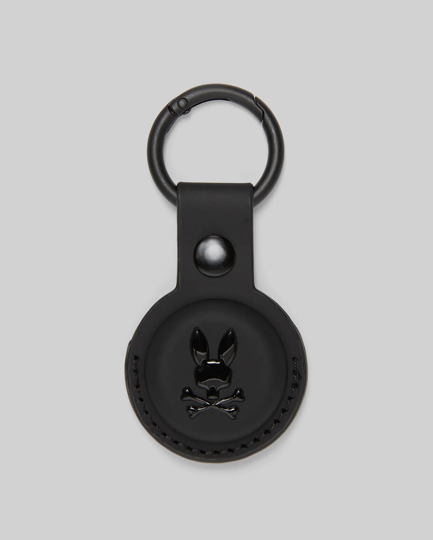 The Psycho Bunny MENS AIR TAG HOLDER - MO1700017 is a round, black leather holder with an embossed bunny logo, snap button, and metal keyring—ideal as a stylish luggage tag.
