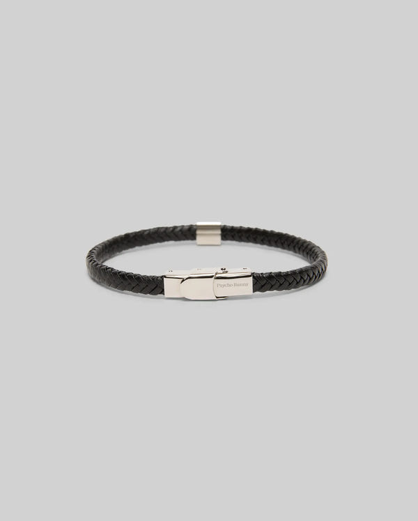 The Psycho Bunny MENS LEATHER BRAIDED BRACELET - MO1700020 features black braided leather, a hinged clasp, and bunny-engraved hardware. It is displayed against a plain light gray background.