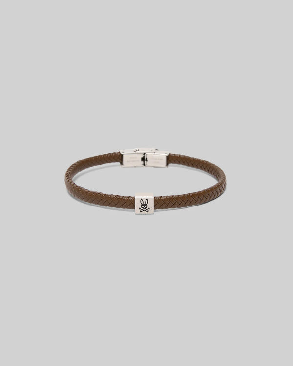 The Psycho Bunny MENS LEATHER BRAIDED BRACELET - MO1700020 features a brown braided leather design, a hinged clasp, and a rectangular hardware charm engraved with the signature bunny and skull-and-crossbones. Displayed on a light gray background.