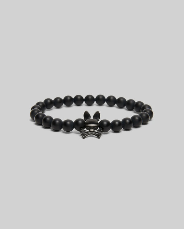 The Psycho Bunny MENS BUNNY BEAD BRACELET (MO1700023) features black beads and a metallic skull-and-crossbones charm with bunny ears—ideal as a subtle fidget or stylish accent—shown on a simple light gray background.