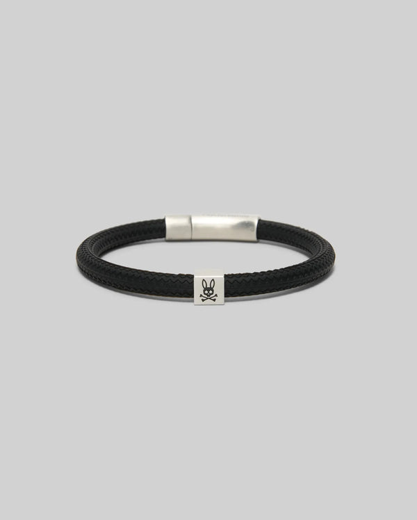 The Psycho Bunny MENS SILICONE BRACELET - MO1700025 features a black braided design with a silver clasp and small skull & crossbones charm, shown on a plain light gray background.