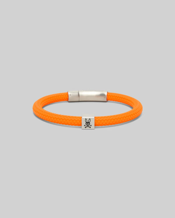 The Psycho Bunny MENS SILICONE BRACELET - MO1700025 features an orange braided design with a silver clasp and a small charm showing a black skull and crossbones with rabbit ears, set against a plain gray background.