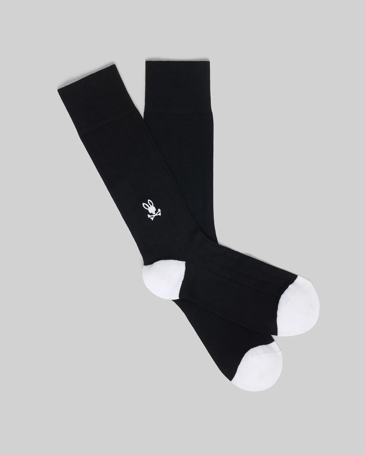The Psycho Bunny MENS 2 PACK DRESS SOCK (MO2200005) features black socks with white toes and heels, and a small white skull and crossbones with bunny ears on the side, displayed overlapping on a plain gray background.