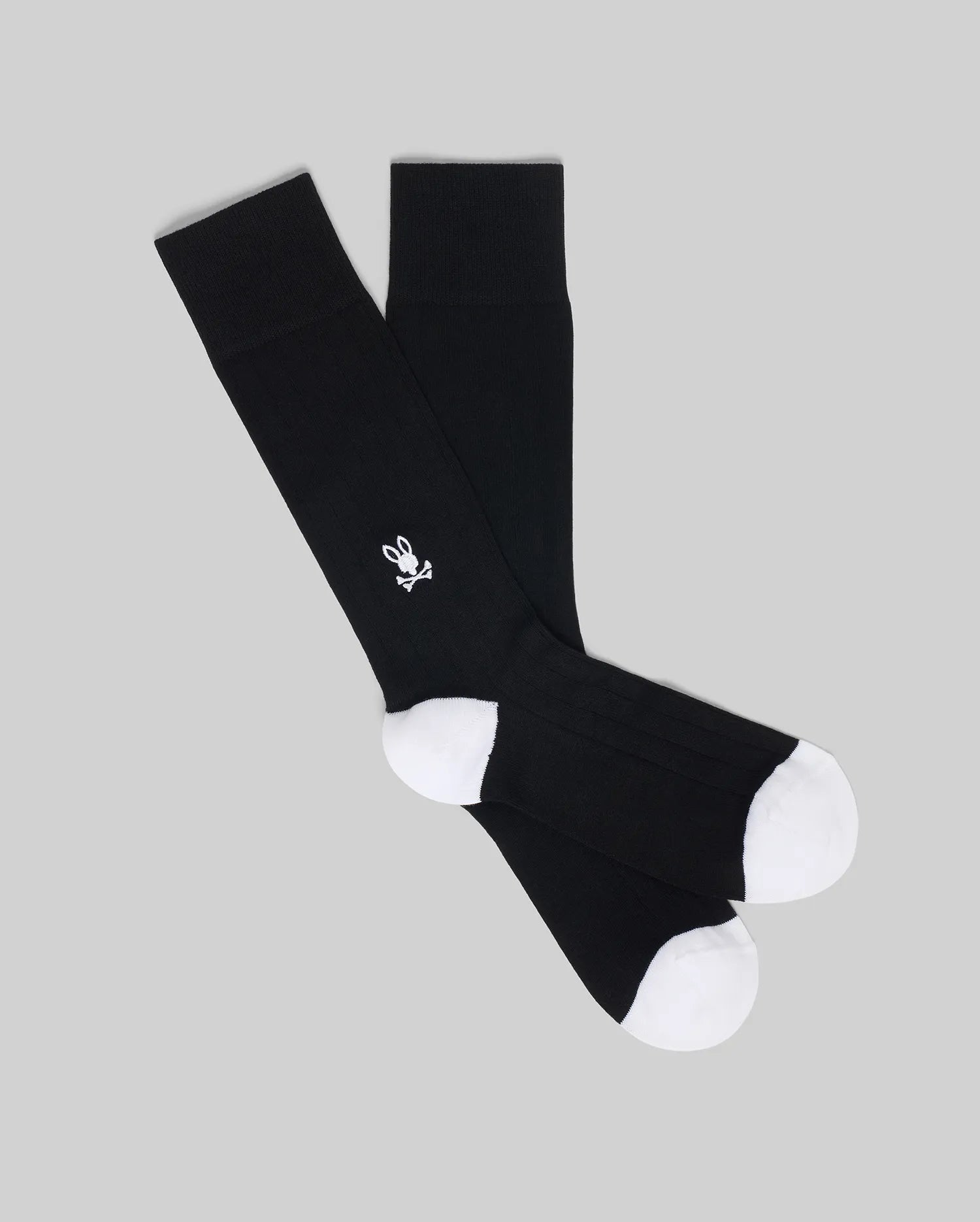 The Psycho Bunny MENS 2 PACK DRESS SOCK (MO2200005) features black socks with white toes and heels, and a small white skull and crossbones with bunny ears on the side, displayed overlapping on a plain gray background.