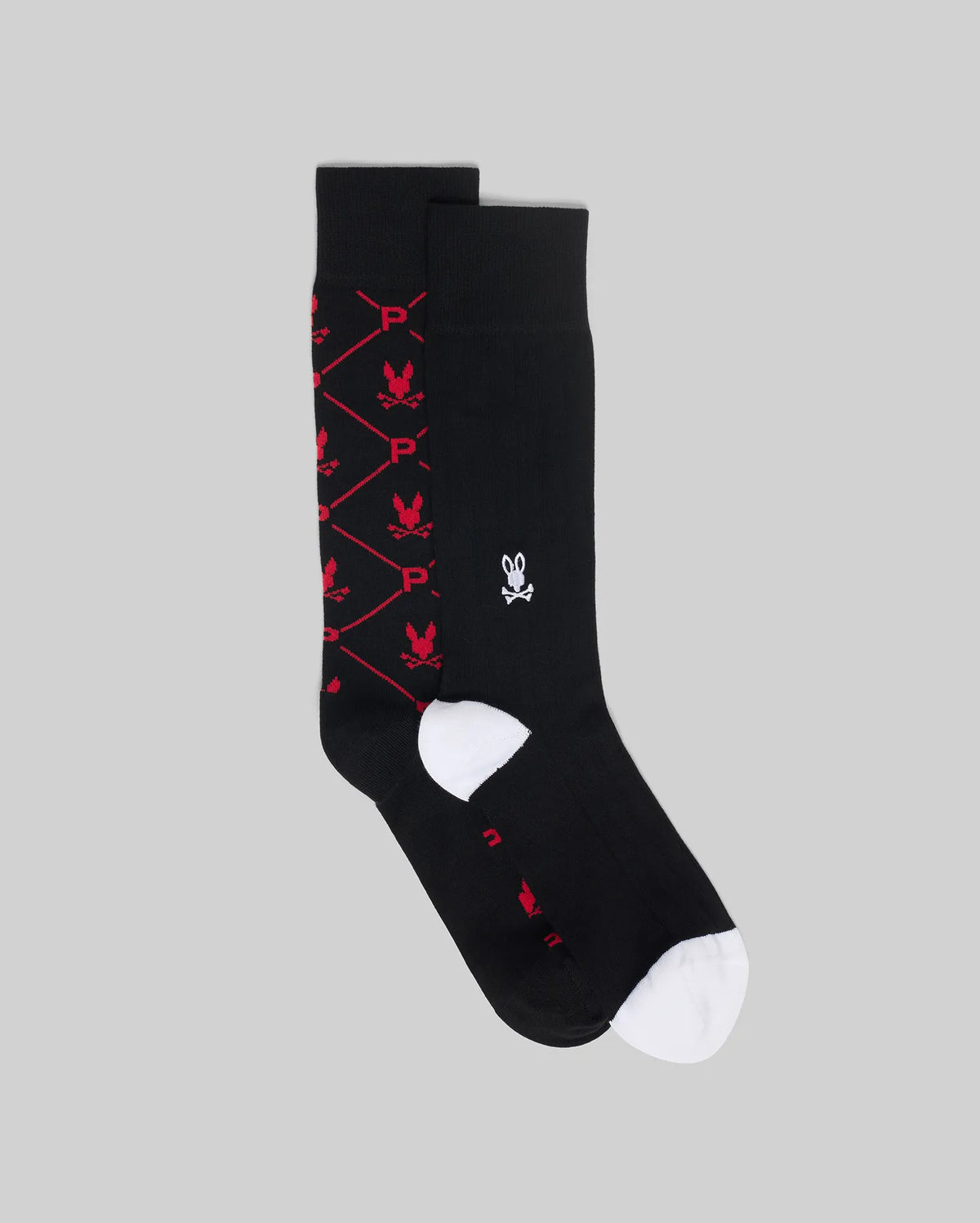 The Psycho Bunny MENS 2 PACK DRESS SOCK (MO2200005) features one black sock with a red diamond bunny “P” design, and another plain black sock with a small white bunny skull logo and white toe, shown on a gray background.