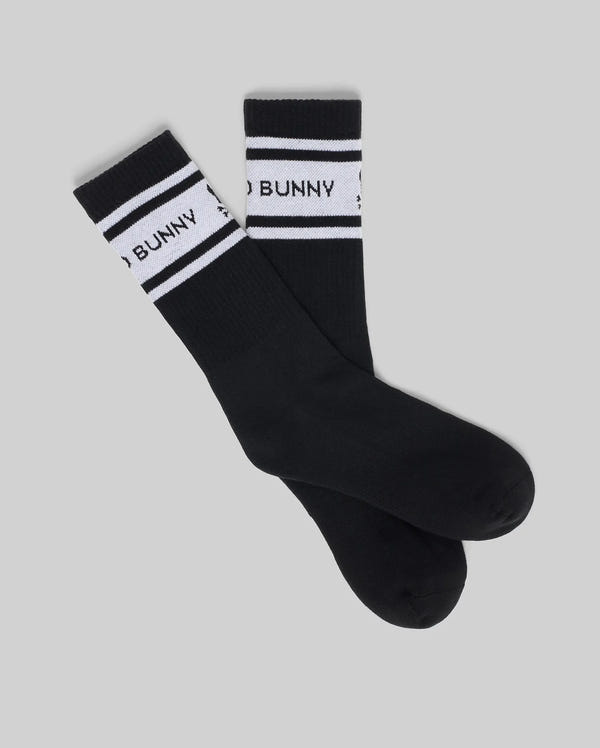 The Psycho Bunny MENS 3 PACK SPORT SOCK (MO2200008) includes black Pima cotton socks with white bands near the cuffs featuring “BUNNY” in black, along with stripes and star details on a plain grey background.