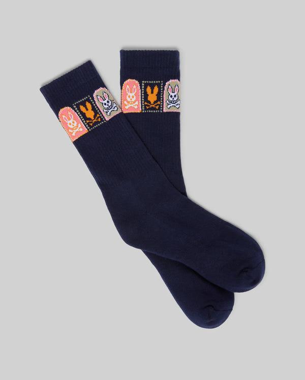 The MENS SUNDEW GRAPHIC SPORT SOCK by Psycho Bunny features navy blue Pima cotton and four colorful cartoon bunny graphics in pink, orange, purple, and blue on a rectangular panel near the top. Displayed on a light gray background.