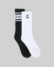 The Psycho Bunny MENS 2 PACK SPORT SOCK (MO2200036) includes one black sock with white stripes and partial text, and one white cotton sock featuring a small black cartoon bunny face, photographed on a gray background.