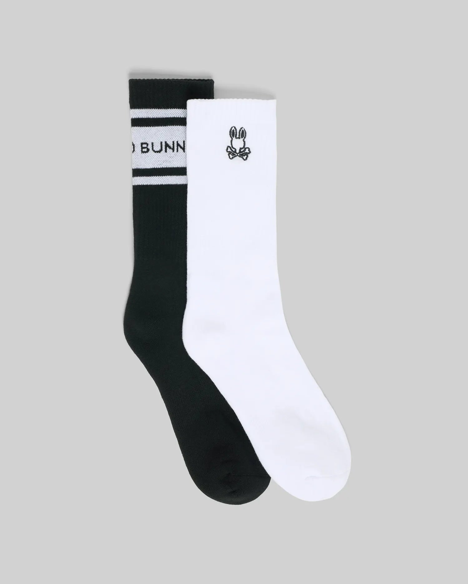 The Psycho Bunny MENS 2 PACK SPORT SOCK (MO2200036) includes one black sock with white stripes and partial text, and one white cotton sock featuring a small black cartoon bunny face, photographed on a gray background.
