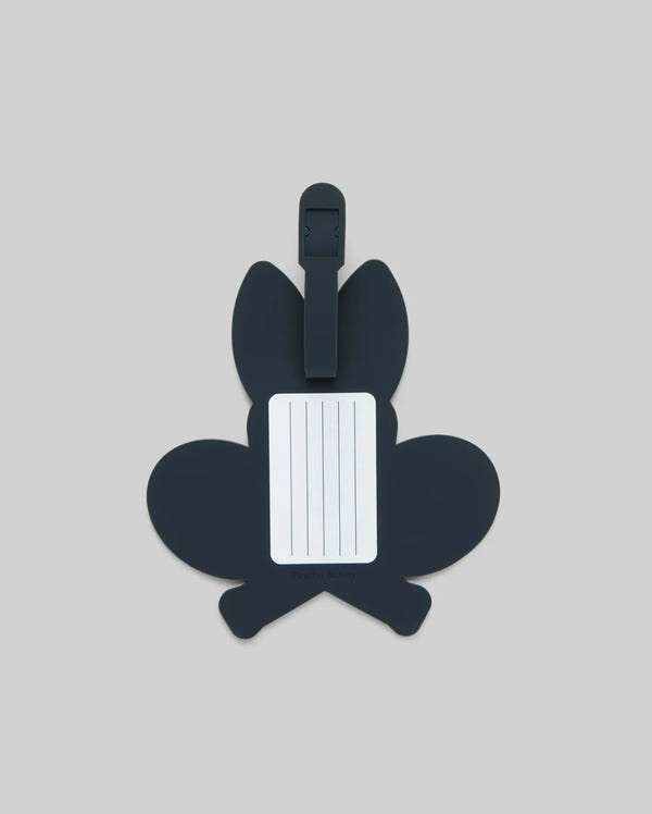 The Psycho Bunny MENS RACKET BUNNY LUGGAGE TAG (MO2400003) features a dark, bunny-shaped silicone design with a white info label and simple strap, shown on a plain light gray background.