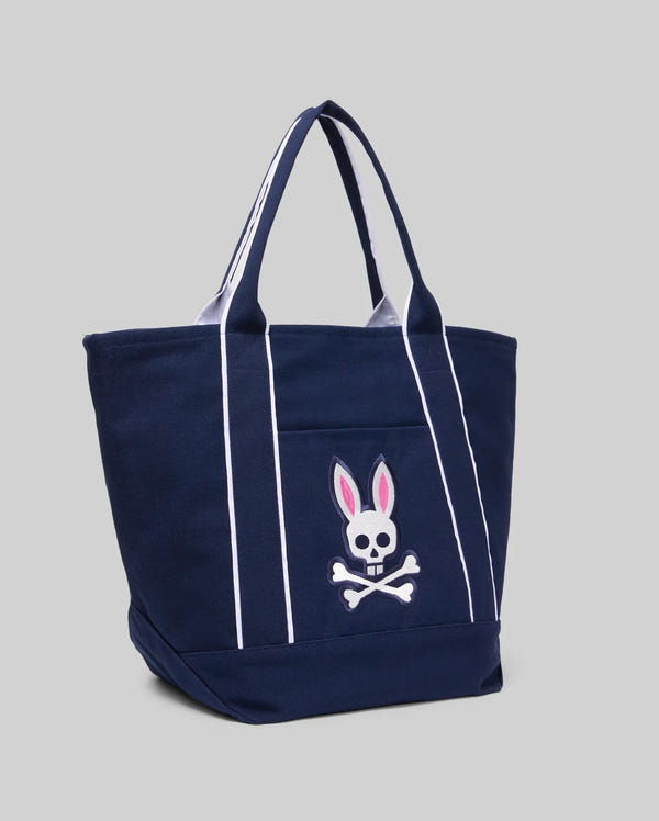 The Psycho Bunny MENS CANVAS TOTE BAG - MO2400016 is a navy blue tote with white piping, two handles, and a front pocket embroidered with a white skull and crossbones topped by pink bunny ears, shown on a plain gray background.