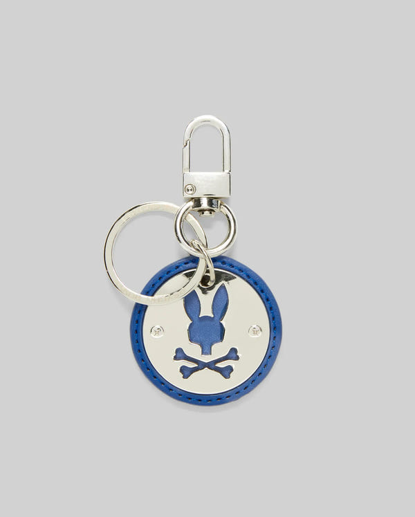 The Psycho Bunny DISC KEYCHAIN - MO2400020 is a round blue leather keychain with a polished metal plate featuring a bunny head above crossed bones, attached to a keyring and lobster clasp. The background is light gray.