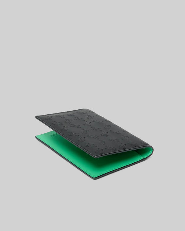 Passport Holder, black with a smooth leather-like texture, featuring an all-over embossed Psycho Bunny skull logo pattern on the exterior and a vibrant green interior.