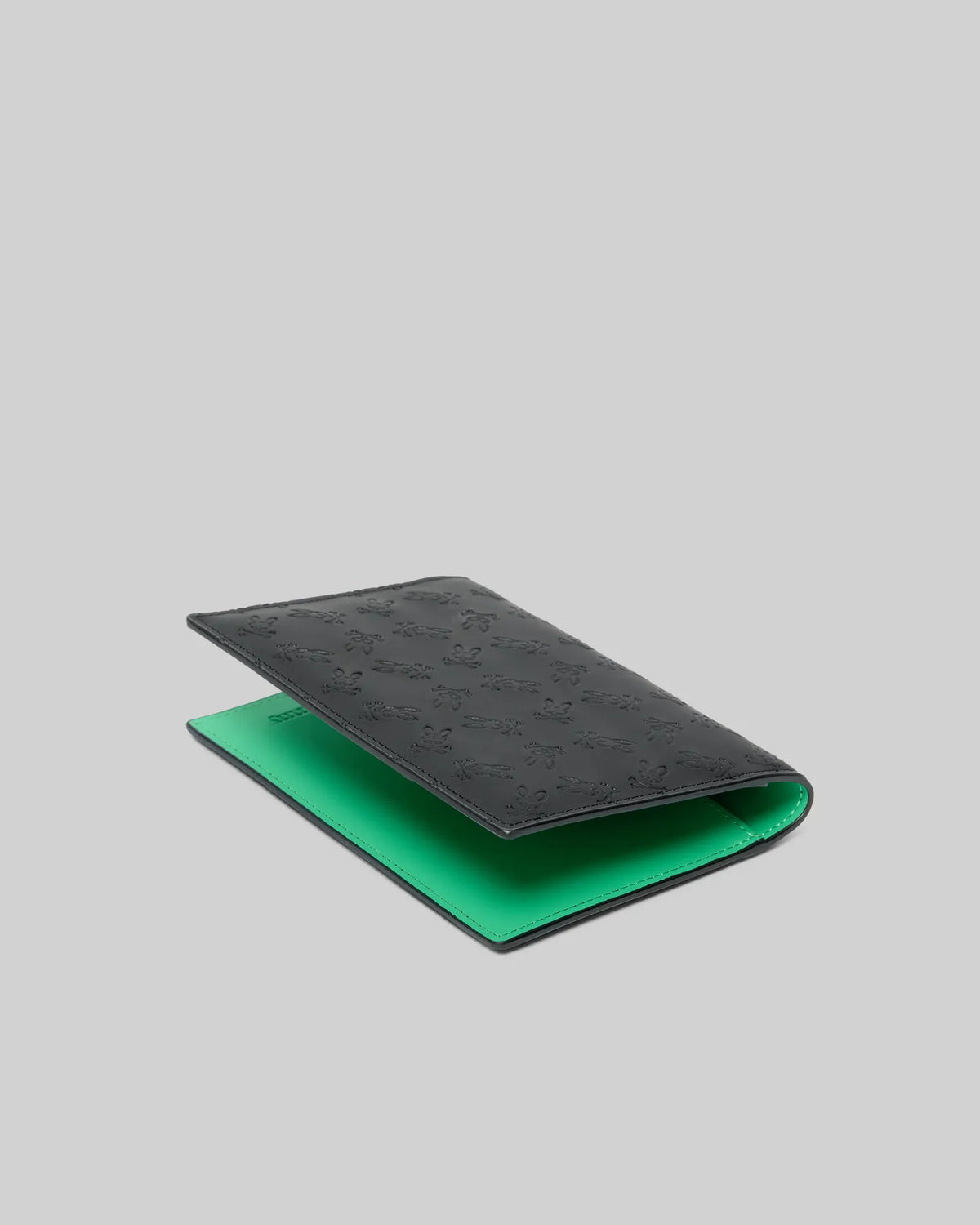 Passport Holder, black with a smooth leather-like texture, featuring an all-over embossed Psycho Bunny skull logo pattern on the exterior and a vibrant green interior.