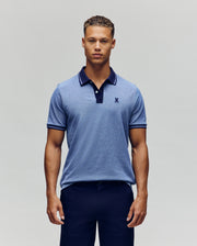 A man stands facing forward, wearing the Psycho Bunny QUINTON TEXTURE PIQUE POLO in light blue with dark trim on the collar and sleeves, paired with dark trousers against a plain, light background.
