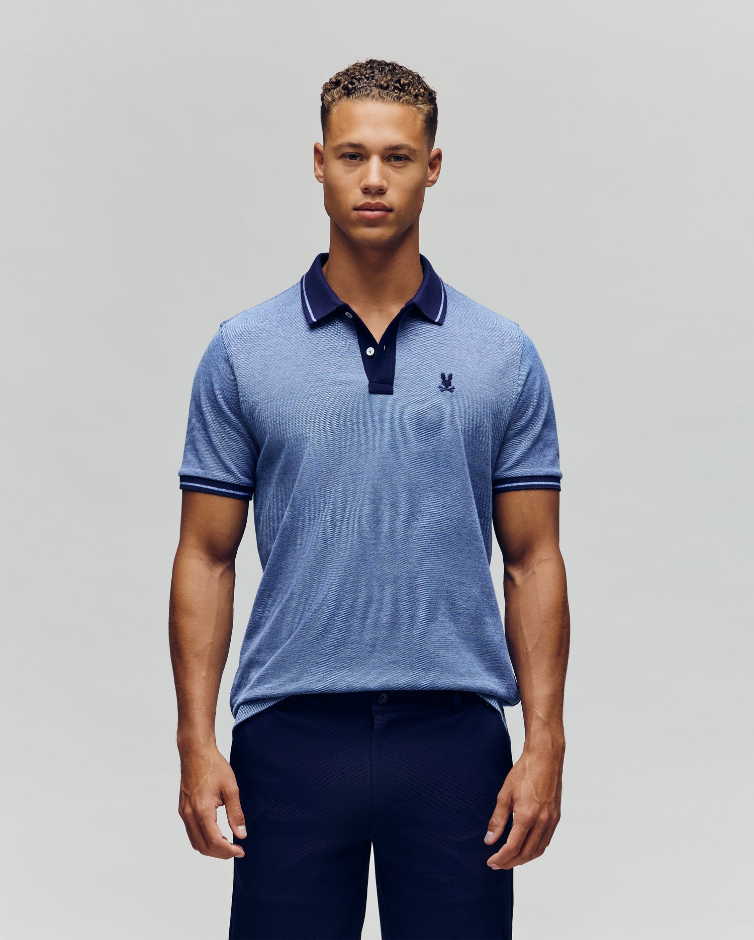 A man stands facing forward, wearing the Psycho Bunny QUINTON TEXTURE PIQUE POLO in light blue with dark trim on the collar and sleeves, paired with dark trousers against a plain, light background.