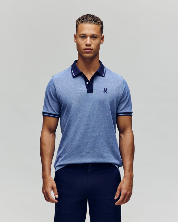 A man stands facing forward, wearing the Psycho Bunny QUINTON TEXTURE PIQUE POLO in light blue with dark trim on the collar and sleeves, paired with dark trousers against a plain, light background.