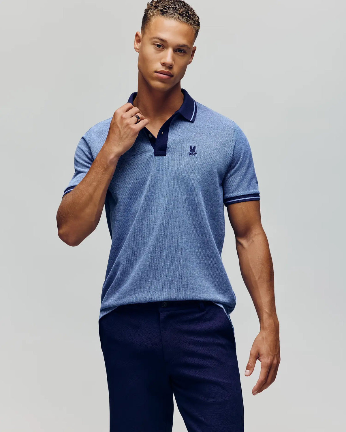 A young man poses confidently against a plain background, wearing the Psycho Bunny QUINTON TEXTURE PIQUE POLO in light blue with dark trim and navy pants, holding the polo's collar as he looks at the camera.