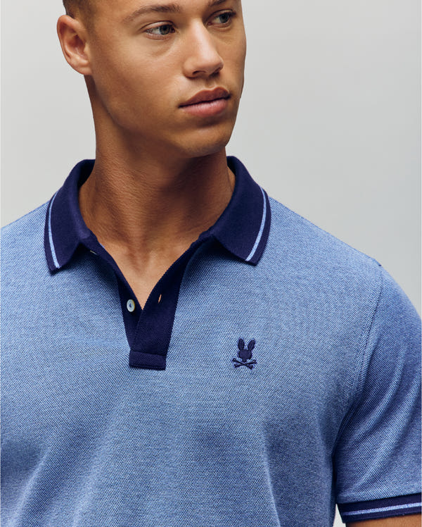 A young man wears the Psycho Bunny QUINTON TEXTURE PIQUE POLO, a light blue shirt with a dark blue collar and small embroidered chest logo, as he looks slightly to the side against a plain background.