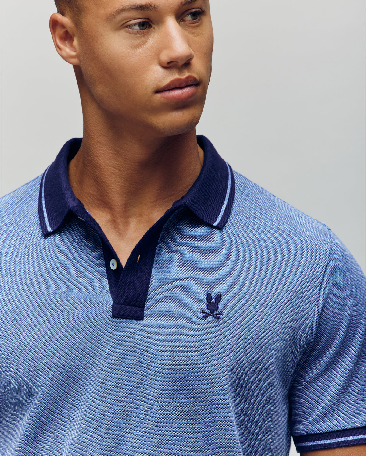 A young man wears the Psycho Bunny QUINTON TEXTURE PIQUE POLO, a light blue shirt with a dark blue collar and small embroidered chest logo, as he looks slightly to the side against a plain background.