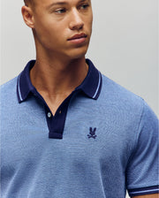 A young man wears the Psycho Bunny QUINTON TEXTURE PIQUE POLO, a light blue shirt with a dark blue collar and small embroidered chest logo, as he looks slightly to the side against a plain background.