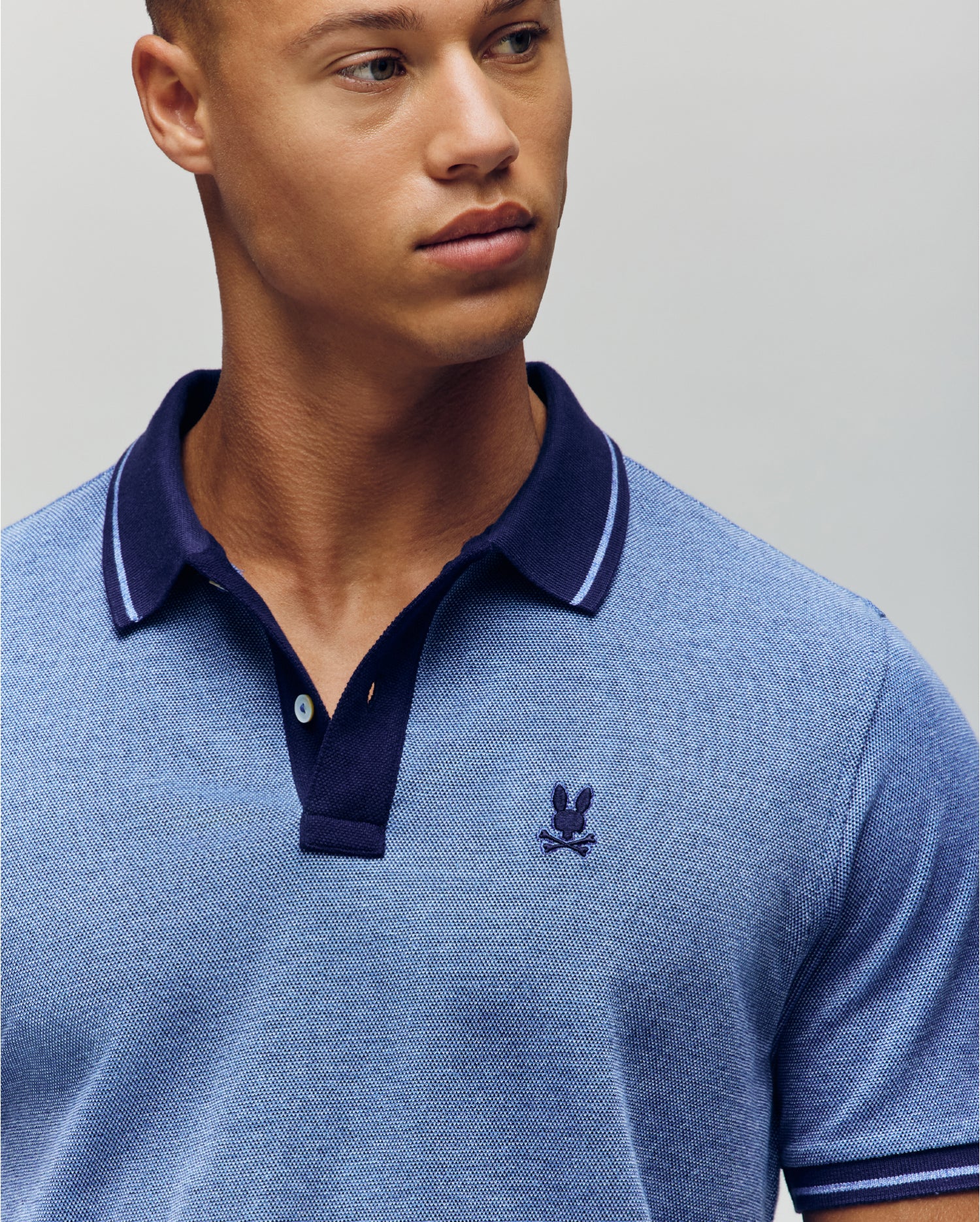 A young man wears the Psycho Bunny QUINTON TEXTURE PIQUE POLO, a light blue shirt with a dark blue collar and small embroidered chest logo, as he looks slightly to the side against a plain background.