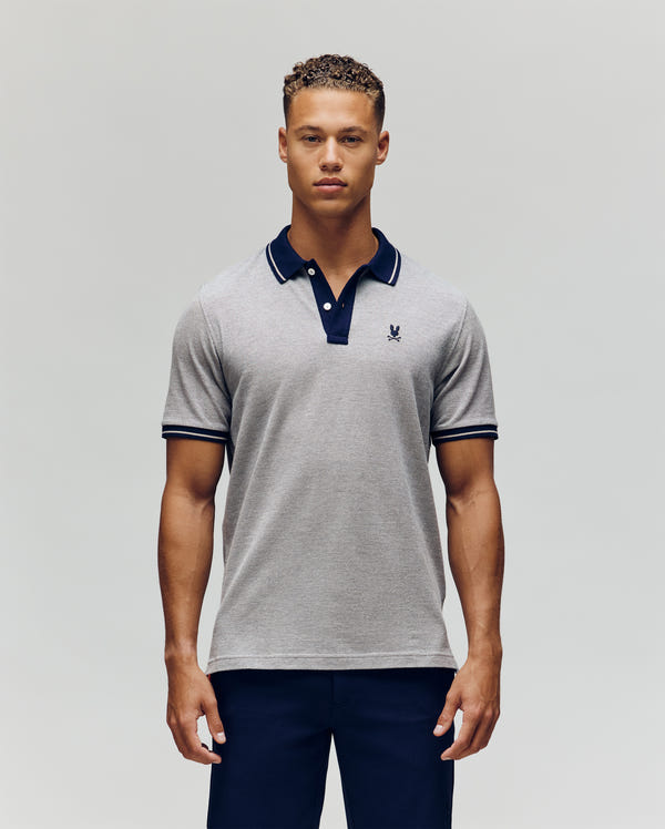 A young man stands facing forward against a plain light background, wearing Psycho Bunny’s QUINTON TEXTURE PIQUE POLO in light grey with dark edging on the collar and sleeves, paired with dark pants.