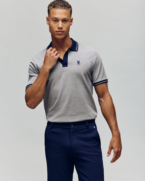 A man wears the Psycho Bunny QUINTON TEXTURE PIQUE POLO—light grey with dark collar and sleeve trim—paired with dark blue pants. He holds the polo’s collar and looks at the camera against a plain background, expression neutral.