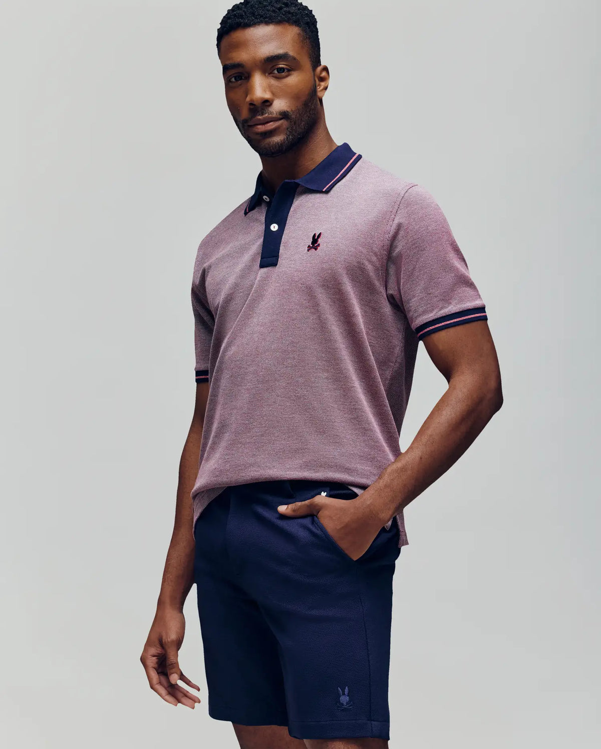 A man stands confidently in a Psycho Bunny QUINTON TEXTURE PIQUE POLO, a short-sleeve light purple shirt with an embroidered logo, and dark navy shorts, set against a plain light gray background.
