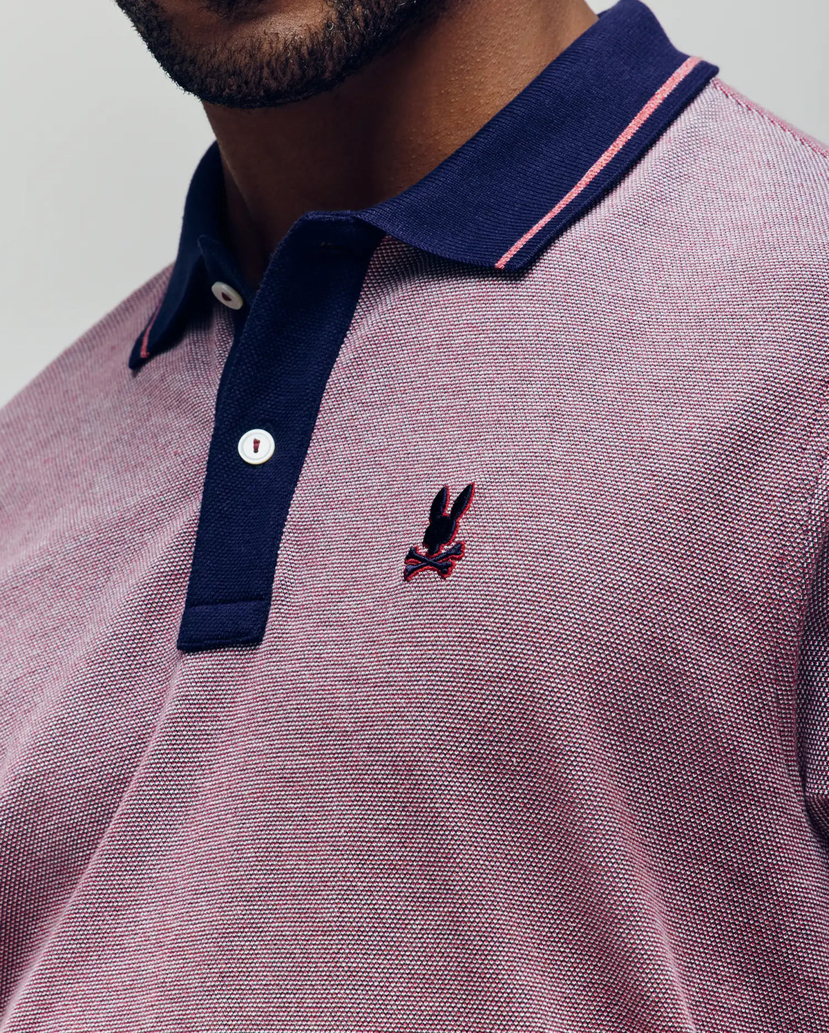 Close-up of a person wearing the Psycho Bunny QUINTON TEXTURE PIQUE POLO in pink and navy, featuring pink accents and an embroidered dark bunny logo on the chest. Only the lower face and upper torso are visible.